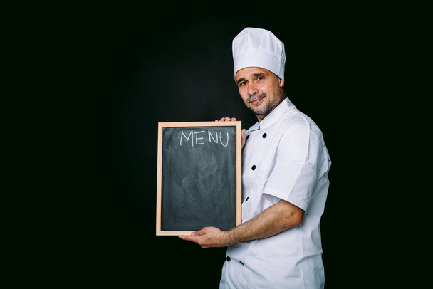 Chef cuisinier vêtu d'une veste et d'un chapeau de cuisine, tenant un tableau noir avec menu, sur fond noir - Photo, image