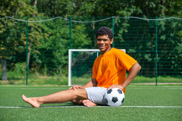 Молодий афроамериканець тримає футбольний м'яч, сидячи на відкритому повітрі проти футбольної мети. - Фото, зображення