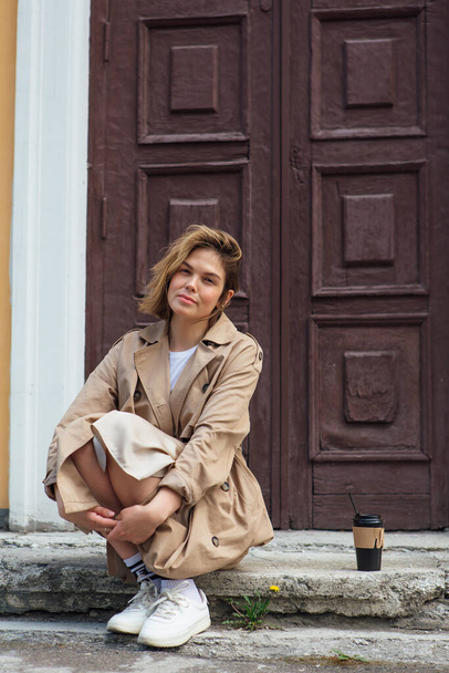 Young smiling millennial woman with wild hair dressed in an autumn coat sitting near the door of an old building with a cup of coffee to go. - Foto, Imagem