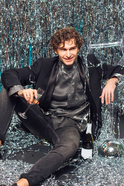 Tall attractive man sitting on the floor over the sparkle background and mirror disco ball holding glass of champagne. Party concept. - Valokuva, kuva