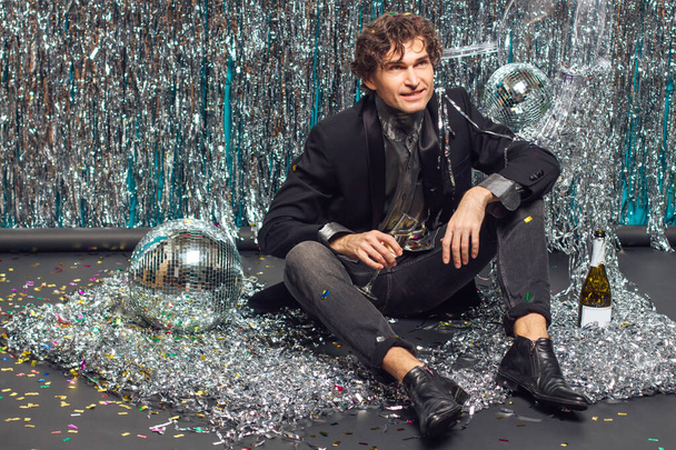 Wysoki atrakcyjny mężczyzna siedzący na podłodze na błyszczącym tle i lustrzana kula dyskotekowa trzymająca kieliszek szampana. Koncepcja partii. - Zdjęcie, obraz