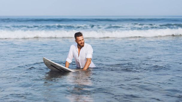 Een jonge Europeaan in een wit shirt zit op een surfplank in de zee en kijkt opzij. - Foto, afbeelding