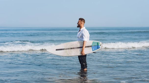 Un giovane europeo in camicia bianca tiene in mano una tavola da surf e sta per fare un giro. - Foto, immagini