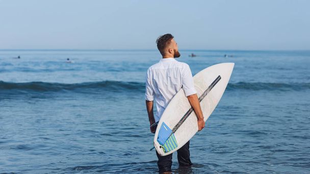 Ένας νεαρός με σανίδα του σερφ με λευκό πουκάμισο πάει στη θάλασσα. Σέρφερ στην παραλία του Μπαλί. Πίσω όψη - Φωτογραφία, εικόνα