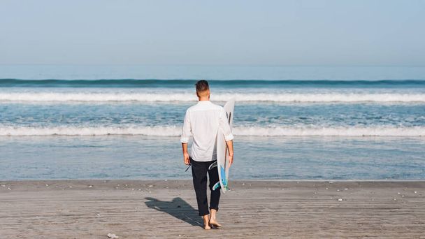 白いシャツを着たサーフボードを着た若い男が海に行く。バリ島のビーチでサーファー。後景 - 写真・画像