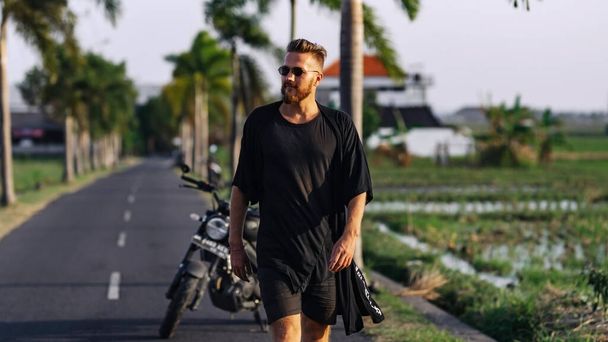 Nuori kaveri tummissa vaatteissa kävelee tietä pitkin moottoripyöränsä edessä tropiikissa. - Valokuva, kuva