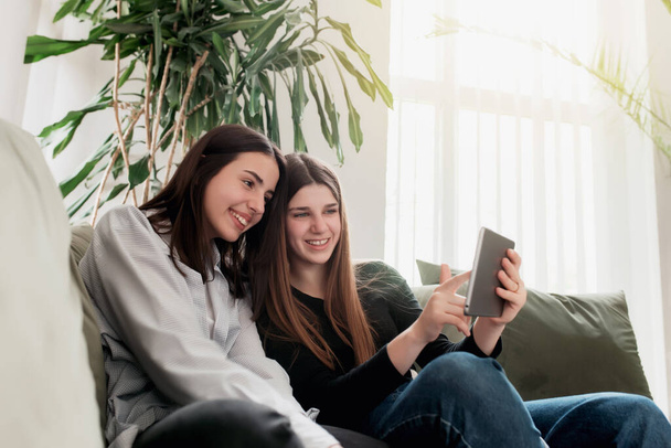 Two teenager sister girls are sitting at green sofa in living room and watching some funny video at tablet, smiling, laughing. Beautiful people, frendship and togetherness concept. Flowerpot on back - Photo, image