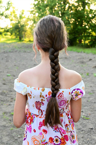 Portrait of beautiful young girl with long hair braided in braid, dressed in summer dress against background of green foliage. Summer outdoor recreation. Rear view - Photo, image