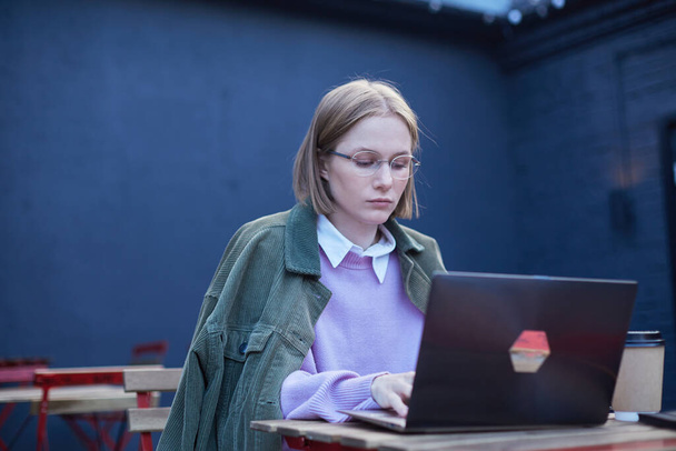 Nachdenkliche junge Geschäftsfrau tippt draußen in Straßencafé auf Laptop - Foto, Bild