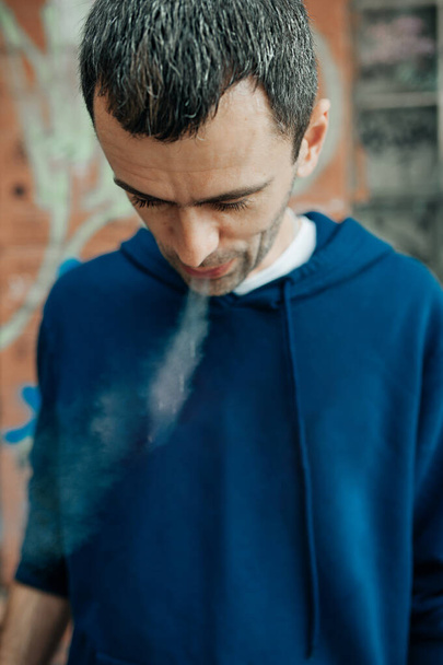 Ένας βίαιος όμορφος άντρας καπνίζει όμορφα με μπλε κουκούλα. Οι άνθρωποι κακές συνήθειες. διαδικασία καπνίσματος. Νέος, γκριζομάλλης, μεσήλικας άντρας βγάζει καπνό από το στόμα του. επιλεκτική εστίαση - Φωτογραφία, εικόνα