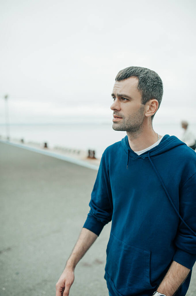 middle-aged man walks on the sea pier. portrait of young man with dark but already partially gray hair in blue hoodie near the sea. urban style content. selective focus - Foto, Bild