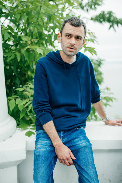 cienki, przystojny młodzieniec w niebieskiej bluzie i dżinsach siedzi na tle zieleni. Portret mężczyzny w wieku 30-40 lat. ukierunkowanie selektywne - Zdjęcie, obraz