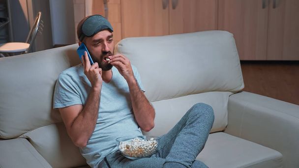 Fehér férfi pihen a kanapén, popcornt eszik, miközben telefonon beszél egy barátjával. - Fotó, kép