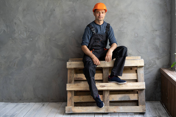 Male worker in work clothes and a construction helmet sits on wooden pallets resting on the background of a concrete wall in the house. The foreman at work sitting by the window. Renovation concept - Photo, image