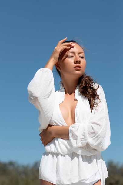 Charmant jong vrouwelijk model in stijlvolle witte blouse onthullende borst staan met de handen op het hoofd en weg te kijken dromerig tegen de blauwe hemel in het zonlicht - Foto, afbeelding