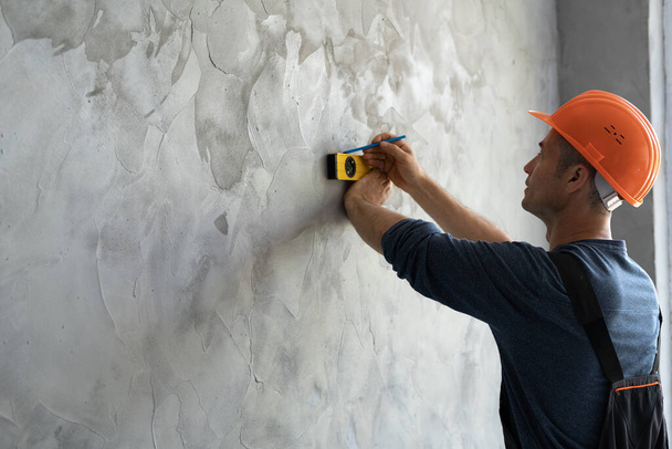 male engineer or architect in a protective helmet measures the level of the wall. A builder in a house is checking a flat concrete wall. Repair and construction concept. Place for text - Photo, image