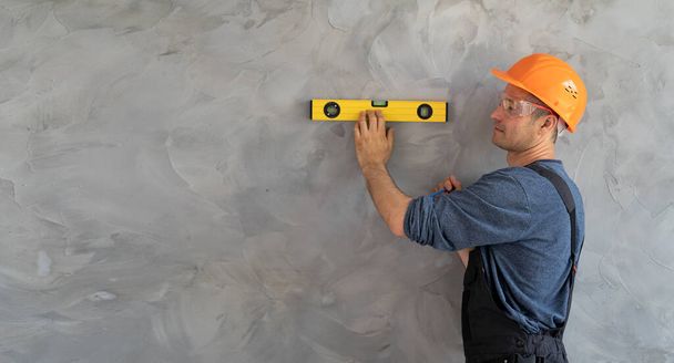 An engineer-architect stands against a wall and measures the level with a ruler. A foreman in a protective helmet checks the work of the builder. House renovation concept. Banner. Place for text - Photo, Image