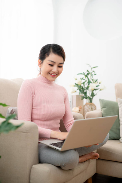 Kaunis iloinen tyttö käyttää hopeinen kannettava tietokone istuessaan sohvalla olohuoneessa kotona. - Valokuva, kuva