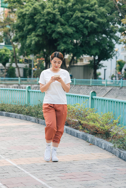 Mujer joven en ropa casual caminando en un parque charlando en su teléfono celular mientras sonríe y espera. - Foto, Imagen
