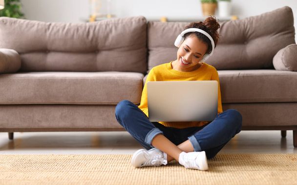 Young happy afro american woman, student or office worker, wearing headphones sitting on floor and listening music, enjoying favourite audio track while working or styding online on laptop at home - Zdjęcie, obraz