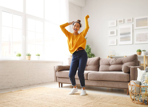 Full length of overjoyed teenage girl in casual wear wearing headphones listening favourite music and dancing while spending carefree time in living room at home. Excited female enjoying leisure - Photo, Image