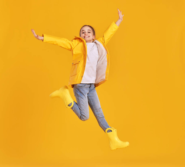 Full body of cheerful energetic preteen girl in yellow raincoat and gumboots leaping with outstretched arms and looking at camera against yellow background - Foto, Imagem