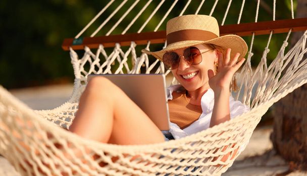 On-line konverzace. Mladá šťastná úspěšná žena nosí slamák mává a usmívá se, zatímco s videohovory na digitálním tabletu, relaxace v houpací síti na tropické písečné pláži za slunečného dne - Fotografie, Obrázek