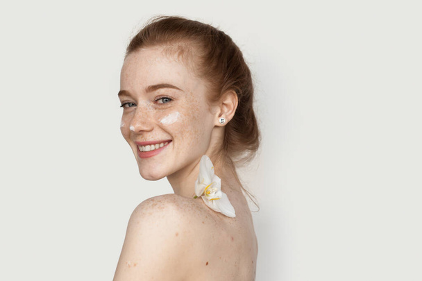 Back view photo of a ginger woman with freckles smiling at camera on a white wall applying anti aging cream on face and holding a flower on shoulder - Фото, изображение