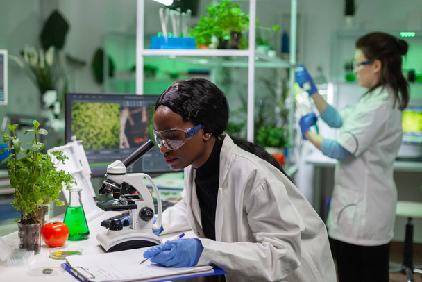 Médico bioquímico africano examinando pruebas químicas con microscopio - Foto, Imagen