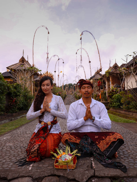 Traditionele Balinese ceremonie. Multicultureel koppel maakt Hindoe religieuze ceremonie met offers. Cultuur en religie. Penjor bamboe decoratie. Blanke vrouw, Balinese man. Penglipuran, Bali - Foto, afbeelding