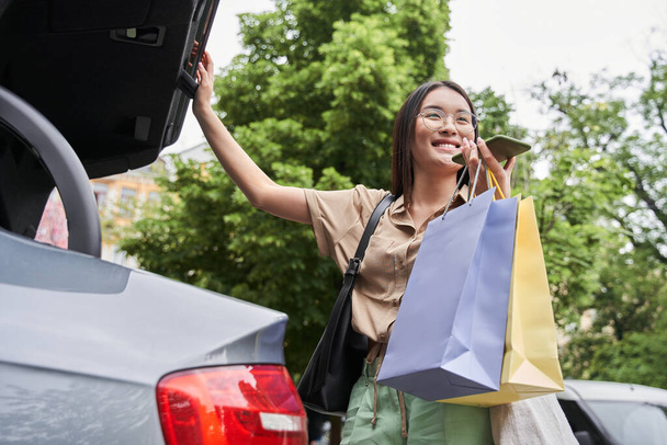 Woman recording voice message and standing near her car while holding shopping bags - Foto, Imagem