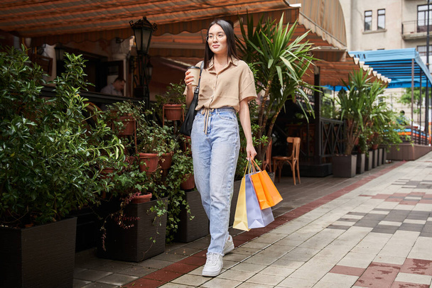 Girl smiling and drinking coffee while stepping with packages from store - Foto, imagen