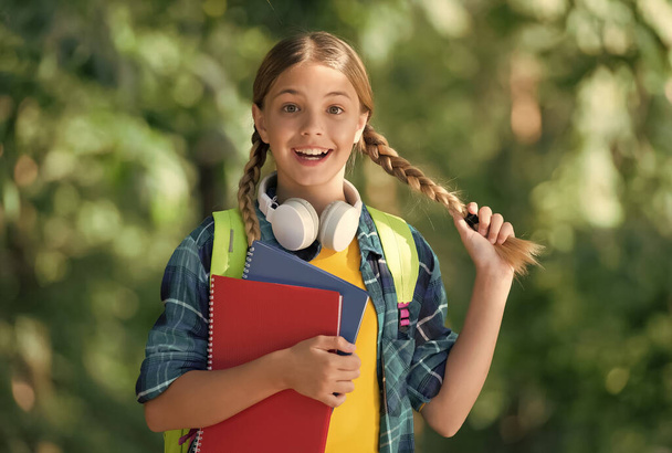 Happy kid with pigtails carry school books for foreign language learning wearing headphones natural landscape, English - Foto, imagen