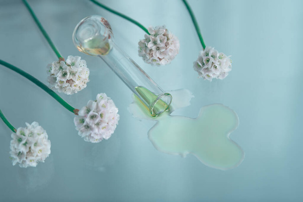 Tubo de vidrio con líquido que gotea en la superficie plateada y flores blancas borrosas. Concepto de esencias y extracciones naturales. - Foto, Imagen