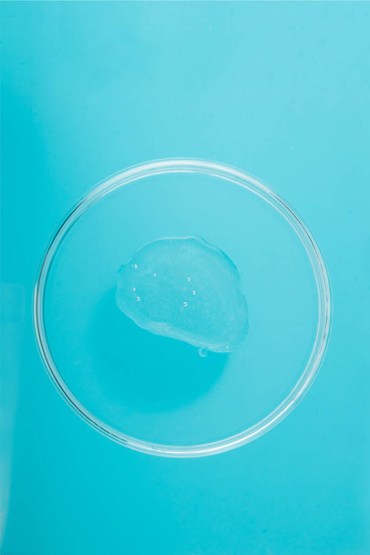 Detailní záběr kapky gelu s třpytkami na kulatém skleněném pohárku a modrým pozadím. Vzorek kosmetického gelu. - Fotografie, Obrázek