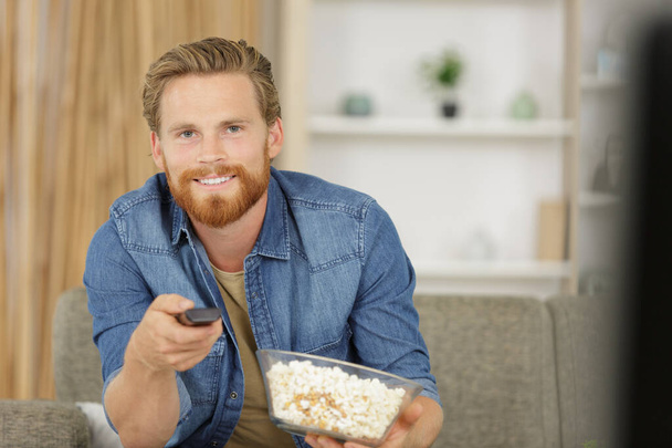 handsome man eats popcorn on sofa in living room - Foto, afbeelding