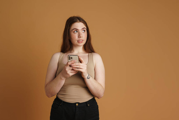Genç kızıl kadın cep telefonu kullanıyor ve bej duvarın üzerinden tek başına bakıyor. - Fotoğraf, Görsel