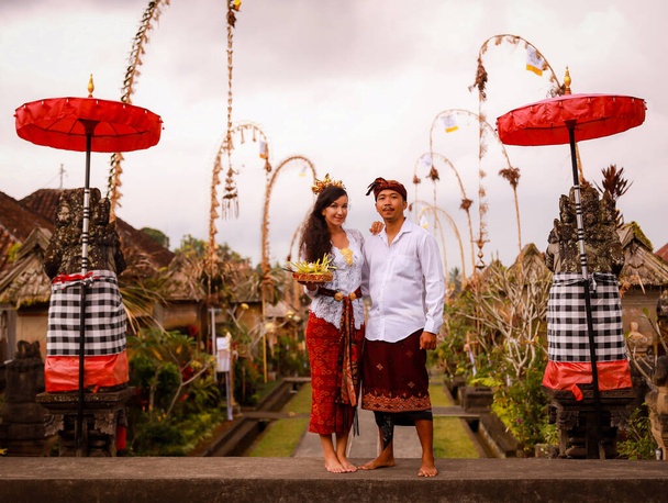 Balinesische Zeremonie. Multikulturelles Paar, das zu einer hinduistischen religiösen Zeremonie mit Opfergaben Gottes geht. Penjor Bambus und Regenschirm Dekoration. Kaukasische Ehefrau und balinesischer Ehemann. Penglipuran, Bali, Indonesien - Foto, Bild