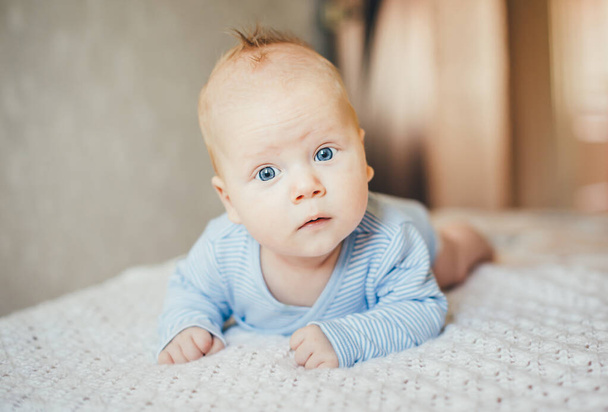 The kid learns to crawl on the bed. He raised his head and looked at the camera in surprise. Funny photo. Blue eyes. - 写真・画像