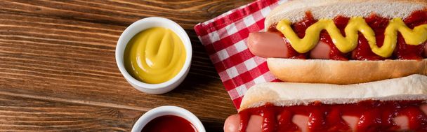 top view of sauce bowls near hot dogs and plaid napkin on wooden table, banner - Photo, Image