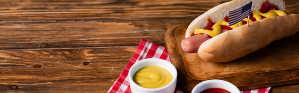tasty hot dog with small usa flag near bowls with mustard and ketchup on wooden table, banner - Photo, Image