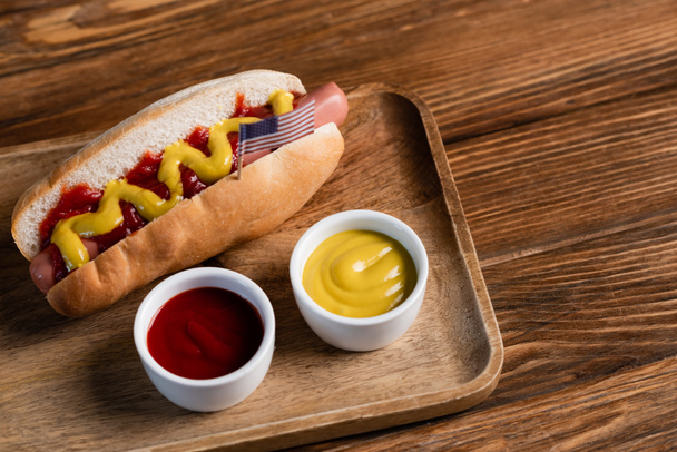 hot dog with small usa flag near bowls with sauces on wooden table - Foto, Bild