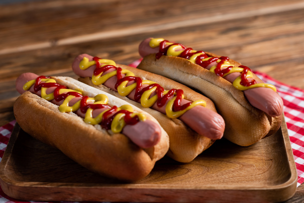 vista de cerca de los perros calientes con mostaza y ketchup cerca de la servilleta a cuadros en la superficie de madera - Foto, Imagen
