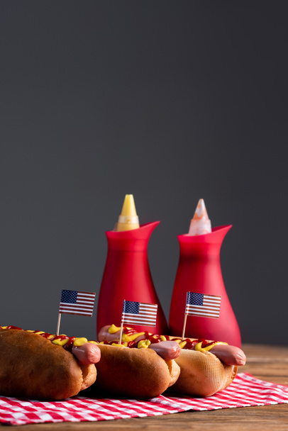 wazig saus flessen en hotdogs met kleine Amerikaanse vlaggen op ruitvormige servet geïsoleerd op grijs - Foto, afbeelding