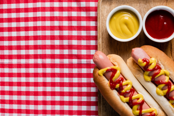 top view of wooden tray with hot dogs and bowls with mustard and ketchup on plaid tablecloth - Fotó, kép