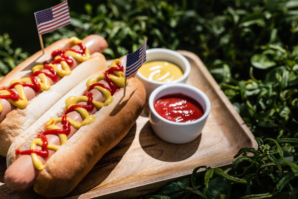 hot dogs with small usa flags near ketchup and mustard on wooden tray and green grass - Photo, Image