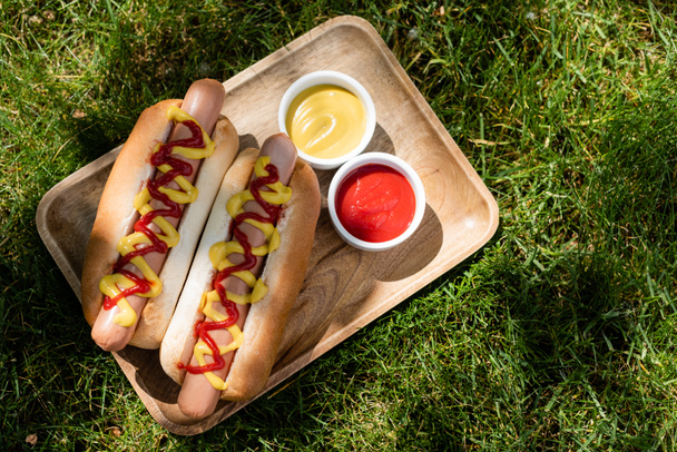 top view of wooden tray with hot dogs, mustard and ketchup on green grass - Zdjęcie, obraz