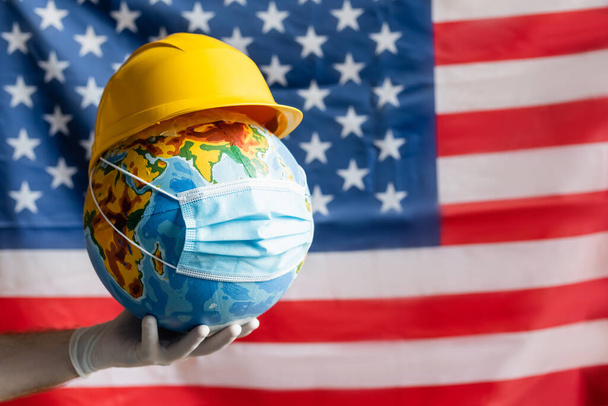cropped άποψη του ανθρώπου στο γάντι λατέξ κρατώντας υδρόγειος σφαίρα με hardhat και ιατρική μάσκα κοντά θολή σημαία ΗΠΑ, έννοια ημέρα εργασίας - Φωτογραφία, εικόνα
