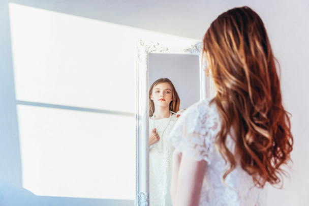 Beauty makeup morning rutine love yourself concept. Young teenage girl looking at reflection in mirror. Young positive woman wearing white dress posing in bright light room against white wall - Foto, afbeelding