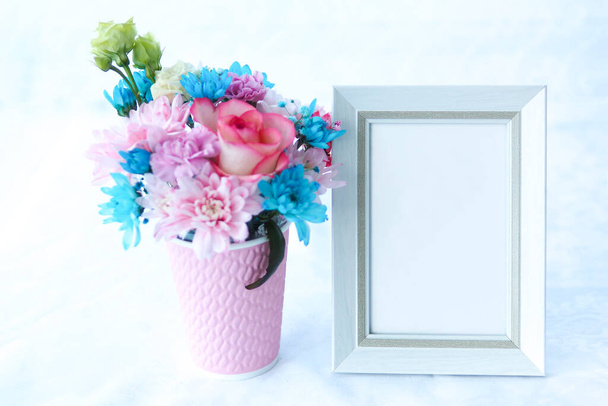 White frame mockup and a bouquet of roses and chrysanthemums in a pink cup on a table with a white tablecloth - Photo, Image